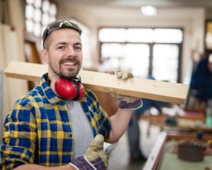 Smiling carpenter carrying a wooden plank in a workshop.